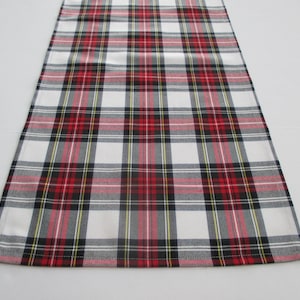 May include: A rectangular plaid table runner in a classic pattern. The design features a white background with a grid of red, black, and yellow lines. The runner is made of fabric and is designed to be placed on a table.