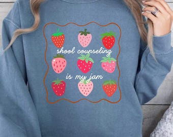 May include: Denim blue sweatshirt with a strawberry graphic and the text "school counseling is my jam". The graphic features nine strawberries in various shades of red and pink, framed by a brown border.