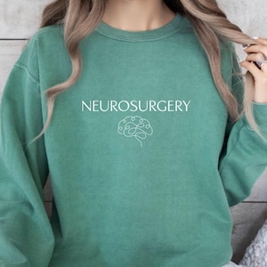 May include: A light green sweatshirt with the word "NEUROSURGERY" printed in white, along with a brain graphic. The sweatshirt is a solid color and has long sleeves. The words "Light Green" are printed at the bottom.
