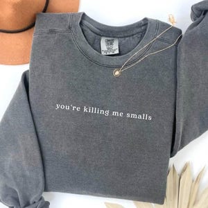 Puede incluir: Una sudadera gris carbón con la frase "you're killing me smalls" en letras blancas. Un collar dorado con un colgante descansa sobre la camisa. Un sombrero marrón es visible en el fondo.