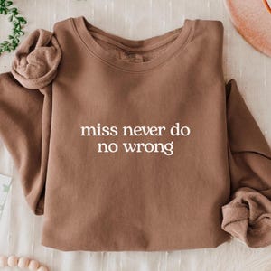 Könnte beinhalten: Ein braunes Sweatshirt mit dem weißen Text "miss never do no wrong". Das Sweatshirt mit Rundhalsausschnitt hat lange Ärmel mit gerafften Details an Schultern und Manschetten. Das Kleidungsstück ist auf einer weißen Oberfläche gefaltet.