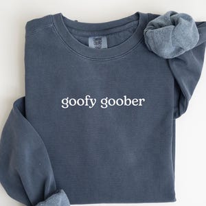 Può includere: Una felpa blu denim con la scritta "goofy goober" in bianco. Le maniche sono arrotolate. La parola "Denim" è stampata in un carattere blu più scuro in basso. La felpa è piegata su una superficie bianca.