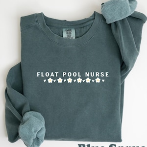 Peut inclure: Un sweat-shirt bleu épicéa avec l'inscription "FLOAT POOL NURSE" en blanc, orné de petites fleurs blanches. Le sweat-shirt a un col rond et des manches longues, avec les manches retroussées. Les mots "Blue Spruce" sont en bas.