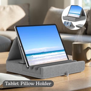 Puede incluir: Un soporte de almohada para tableta gris con una tableta que muestra una escena de playa. El soporte tiene una ranura para la tableta y un pequeño botón. El texto "Tablet Pillow Holder" está en la parte inferior.