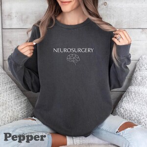May include: A dark gray sweatshirt with the word "NEUROSURGERY" in white, above a white line drawing of a brain. The word "Pepper" is at the bottom of the image.