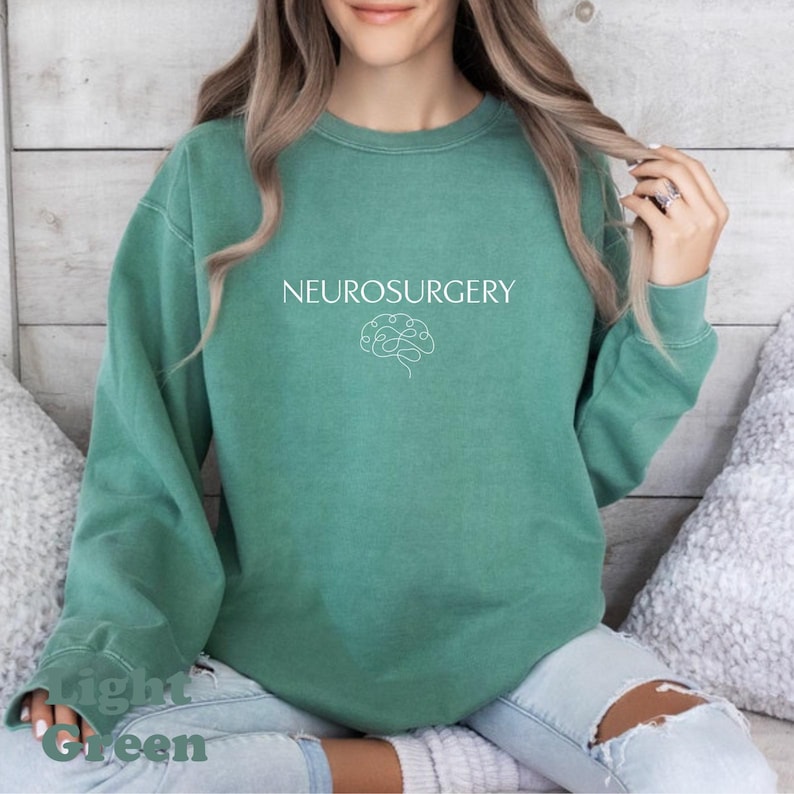 May include: A light green sweatshirt with the word "NEUROSURGERY" printed in white, along with a brain graphic. The sweatshirt is a solid color and has long sleeves. The words "Light Green" are printed at the bottom.