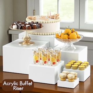 May include: A tiered display of white acrylic buffet risers. The display holds a cake, chocolates, fruit, and drinks. The text "Acrylic Buffet Riser" is at the bottom. The cake has candles and decorative frosting.