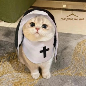 以下が含まれることがあります： 黒いベールと黒い十字架が付いた白いよだれかけを含む、尼僧の習慣を着た明るい色の猫。猫は大きく表現力豊かな目を持ち、模様のあるラグの上に座っています。「MR Print Tee Hub」というテキストが背景に表示されています。
