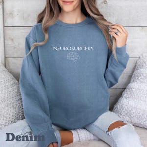 May include: Denim blue sweatshirt with the word "NEUROSURGERY" in white, above a white brain graphic. The word "Denim" is printed at the bottom of the sweatshirt. The sweatshirt is a crew neck style.
