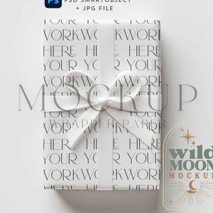 May include: White gift box with a white ribbon and bow. The box is wrapped in paper with a repeating pattern of the words "Your Work Here" in a minimalist font. The box is sitting on a white surface. The text "Wild Moon Mockup" is printed on a label in the lower right corner of the image.