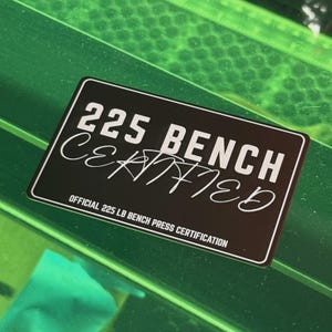 Peut inclure: Un autocollant rectangulaire noir avec le texte blanc « 225 BENCH CERTIFIED ». L'autocollant comprend également le texte « OFFICIAL 225 LB BENCH PRESS CERTIFICATION ». L'autocollant est sur une surface verte.