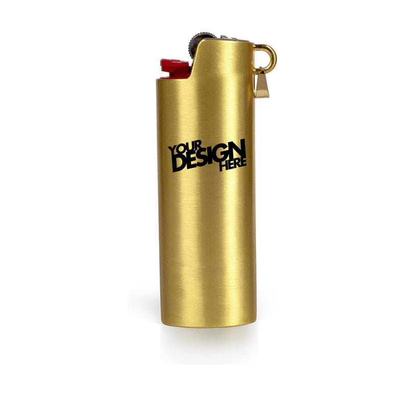 May include: A gold-colored lighter with a red ignition button. The lighter has the text "YOUR DESIGN HERE" printed in black. The lighter is cylindrical with a small loop on the side.