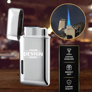 May include: A silver lighter with the top open, displaying a blue flame. The lighter has the text "YOUR DESIGN HERE" on the front. A black sign with gold trim displays "CUSTOM ENGRAVED", "PERFECT GIFT", and "LASTING QUALITY".