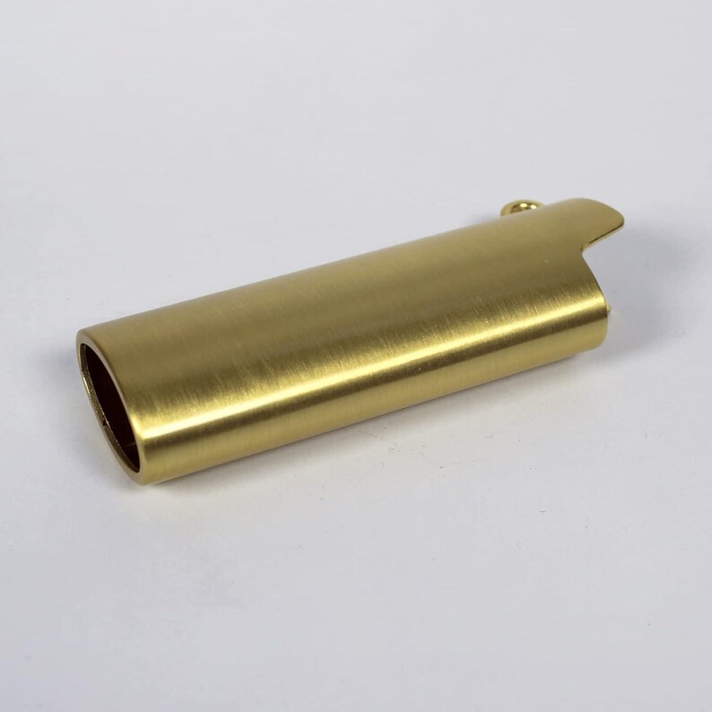 May include: A brass-colored lighter case with a brushed finish. The cylindrical case has a rounded oval opening on one end and a curved, notched top on the other. A small loop is attached to the top. The case is designed to hold a standard-sized lighter.