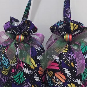 May include: Two purple fabric bags with butterfly patterns and colorful accents. Each bag has a pink and green ribbon tied around it with a colorful bead in the center.