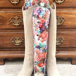 May include: A pair of beige cowboy boots with a floral print design. The boots are standing in front of a wooden dresser. The floral print features a variety of colors, including red, pink, and blue.