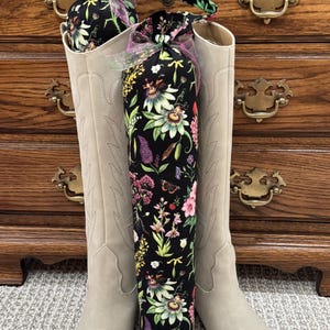 May include: A pair of tall beige boots with pointed toes and stitched detailing, each containing a cylindrical fabric boot shaper. The shapers are made of black fabric featuring a colorful botanical print of flowers and butterflies, tied with a sheer pink ribbon.