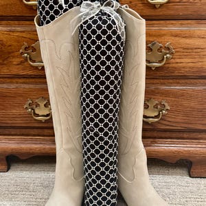 May include: A pair of beige suede cowboy boots with decorative stitching, holding two black and white patterned boot shapers. The shapers are tied with white ribbons and embellished with a small jewel.