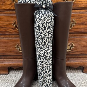 May include: A pair of tall dark brown leather boots with smooth shafts and rounded toes. Inside each boot is a tall, cylindrical fabric boot shaper featuring a black and white leaf pattern, tied at the top with a thin black ribbon.