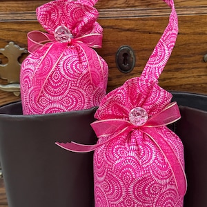May include: Two bright pink fabric boot shapers featuring a white fan-like pattern. Each shaper has a matching pink fabric handle, a sheer pink ribbon tied in a bow, and a clear faceted bead accent. The shapers are inserted into dark brown boots.