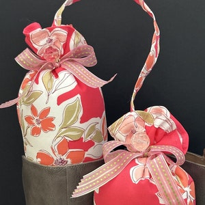 May include: Two pink and white floral fabric bags with a pink ribbon and clear button closure. The bags have a loop handle and are hanging from a brown leather boot.
