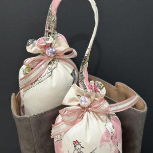 May include: Two fabric sachets with pink ribbon bows and pearl accents. The sachets are filled with a fragrant substance and are designed to be placed in shoes or other enclosed spaces to freshen the air. The sachets are hanging from a loop on a brown boot.
