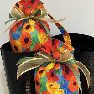 May include: Two fabric bags with a floral print of red poppies, yellow roses, and green leaves on a blue background. The bags are tied with gold and green ribbon and have a red bead accent.