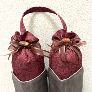 May include: A pair of burgundy shoe bags with floral patterns and brown ribbon ties. Each bag has a colorful ceramic button.