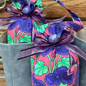 May include: Two fabric gift bags with a floral pattern of purple flowers, green leaves, and pink background. Each bag is tied with a purple ribbon with gold trim and a purple bead. The bags are inside a gray container.