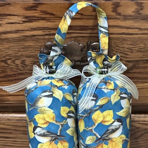 May include: Two cylindrical fabric door stoppers with a blue background, yellow leaves, and bird pattern. Each stopper is tied with a sheer ribbon and has a matching fabric handle. The stoppers are hanging on a decorative metal hook.