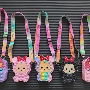 May include: Five colourful crossbody bags with cartoon character designs. Each bag features a different character, including a unicorn, a bear, and a mouse. The bags have rainbow-coloured pop-it style fronts and matching straps with a repeating pattern.