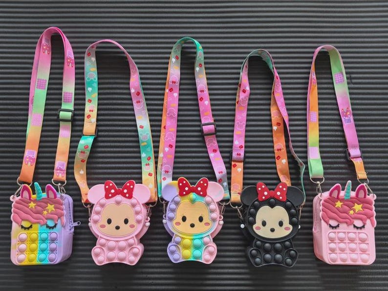 May include: Five colourful crossbody bags with cartoon character designs. Each bag features a different character, including a unicorn, a bear, and a mouse. The bags have rainbow-coloured pop-it style fronts and matching straps with a repeating pattern.