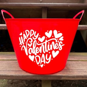 Puede incluir: Un cubo rojo brillante con dos asas, con el texto blanco "Happy Valentine's Day" y motivos de corazón. El cubo está hecho de un material brillante, ideal para regalos o decoraciones de San Valentín.