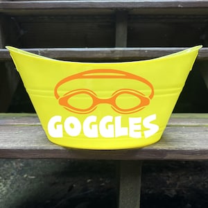 May include: A bright yellow bucket with an orange graphic of swim goggles and the word "GOGGLES" in white. The bucket is made of a shiny material and is sitting on a wooden surface.