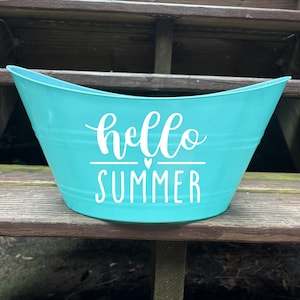Puede incluir: Una tina de metal turquesa con un diseño curvo. Las palabras "hello SUMMER" están escritas en blanco en la parte delantera. La tina está sobre una superficie de madera.