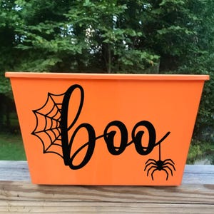 Puede incluir: Un cubo rectangular naranja de Halloween con una telaraña negra, la palabra "boo" en fuente cursiva y una araña colgante. El cubo es de plástico y está sobre una superficie de madera.