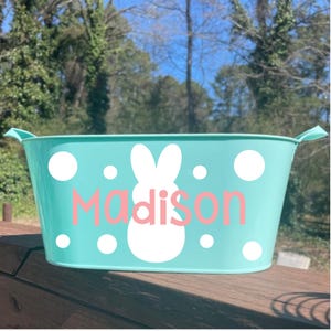 May include: A turquoise metal Easter basket with white polka dots and a white bunny silhouette. The name "Madison" is printed in pink. The basket has two handles and is outdoors.