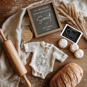May include: A wooden rolling pin, a loaf of bread, and a baby onesie with the text "Baby Smith" are arranged on a wooden surface. A framed sign reads "We are baking something extra sweet December 2025". There are also cookies and an ultrasound image.