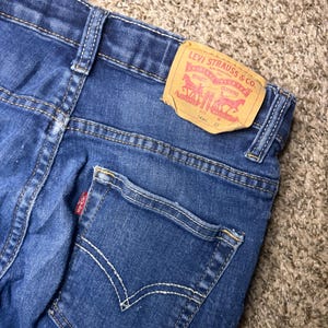 Może przedstawiać: Zbliżenie niebieskich dżinsów z tylną kieszenią i czerwoną metką Levi Strauss & Co. Na metce widnieje nazwa marki, logo i informacje o rozmiarze. Dżinsy mają białe szwy.