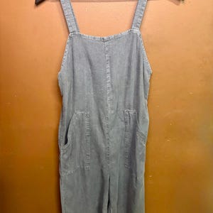 May include: A gray jumpsuit with adjustable shoulder straps and two front pockets hangs on a black hanger. The garment is made of a soft, textured fabric and has a relaxed fit. The jumpsuit is a solid color and appears to be a casual style.