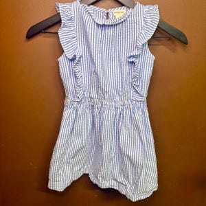 Puede incluir: Vestido a rayas azules y blancas con mangas con volantes y cintura fruncida. El vestido sin mangas cuelga de una percha negra. El cuello del vestido tiene la palabra "crewcuts".
