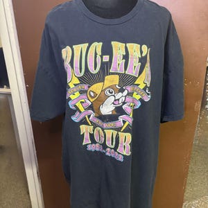 Könnte beinhalten: Schwarzes T-Shirt mit einem farbenfrohen Aufdruck eines Bibers mit Cowboyhut, dem Text "Buc-ee's Tour" und den Jahren "1982-2022". Das Design beinhaltet Blitze und Bänder.