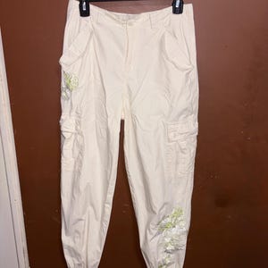 May include: Cream cargo trousers with multiple pockets and a button closure. The trousers feature floral embroidery in shades of green and white near the ankles. The trousers are hanging on a black hanger.