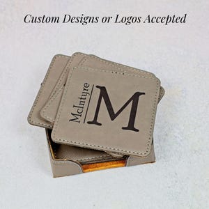 May include: A set of square, gray coasters with the name "McIntyre" and a large letter "M" printed in black. The coasters are stacked in a matching holder. The text "Custom Designs or Logos Accepted" is at the top.