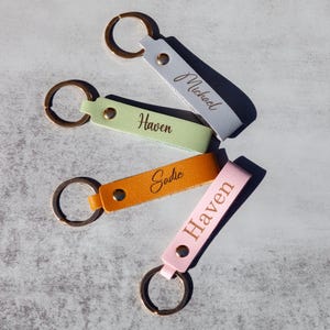 May include: Four leather keychains with gold key rings. The keychains are in different colors: light blue, light green, orange, and pink. Each keychain has a name engraved on it: Michael, Haven, Sadie, and Haven.