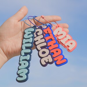 May include: A hand holds four colorful name tags against a blue sky. The tags read "WILLIAM", "CHLOE", "ETHAN", and "MIA" in a bubble letter style. Each tag has a different color scheme, with a matching lanyard.