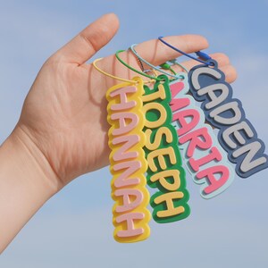 May include: A collection of colorful name tags with the names "Hannah", "Joseph", "Maria", and "Caden" in a playful font. Each tag has a different color scheme, with yellow, green, pink, and blue. The tags are held against a blue sky background.