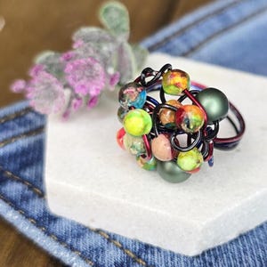 May include: A handmade ring with a cluster of colorful beads. The beads are various sizes and colors, including green, yellow, red, and blue. The ring's wire is black, red, and blue. The ring is on a white surface with a denim background.
