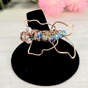 May include: A copper wire ring in a butterfly design, adorned with blue, clear, and pink beads. The ring is displayed on a black stand, showcasing the intricate wirework and colorful accents. The ring is a unique piece of jewelry.