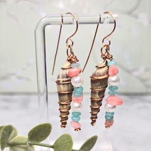 May include: A pair of handcrafted earrings featuring spiral seashells, copper wire, and colorful beads. The earrings have a copper hook and are adorned with pink, white, and turquoise beads, creating a coastal theme.